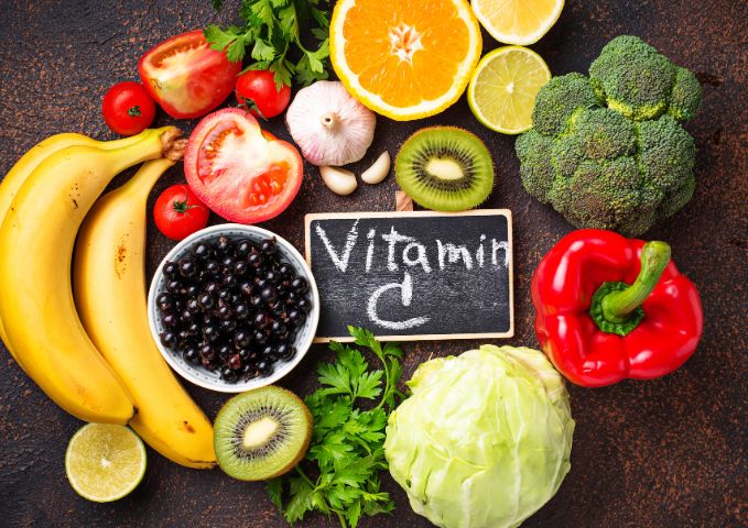 receitas ricas em vitamina C: Várias frutas como banana, laranja kiwi, limão; legumes: brócolis, repolho, pimentão vermelho, tomate. Todos juntos em cima de uma mesa.