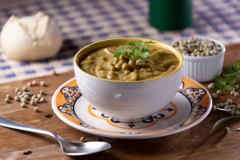 caldo de ervilha em uma tigela com uma colher ao lado e um pão francês ao fundo.