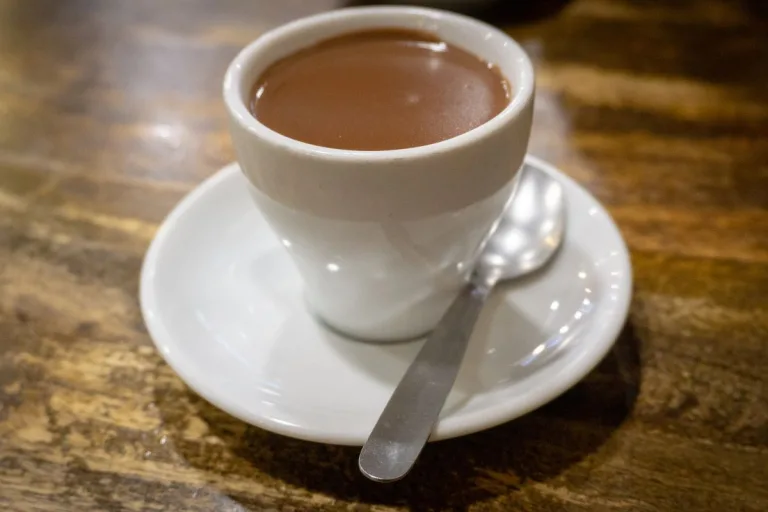 chocolate quente cremoso em uma xícara branca, pires e uma colher ao lado.