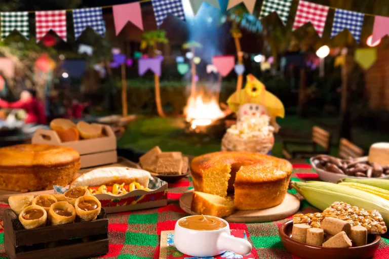 receitas doces de festa junina: mesa lindamente decorada com toalha xadrez, espantalho e vários doces típicos de festa junina. Ao fundo uma festa junina com fogueira.
