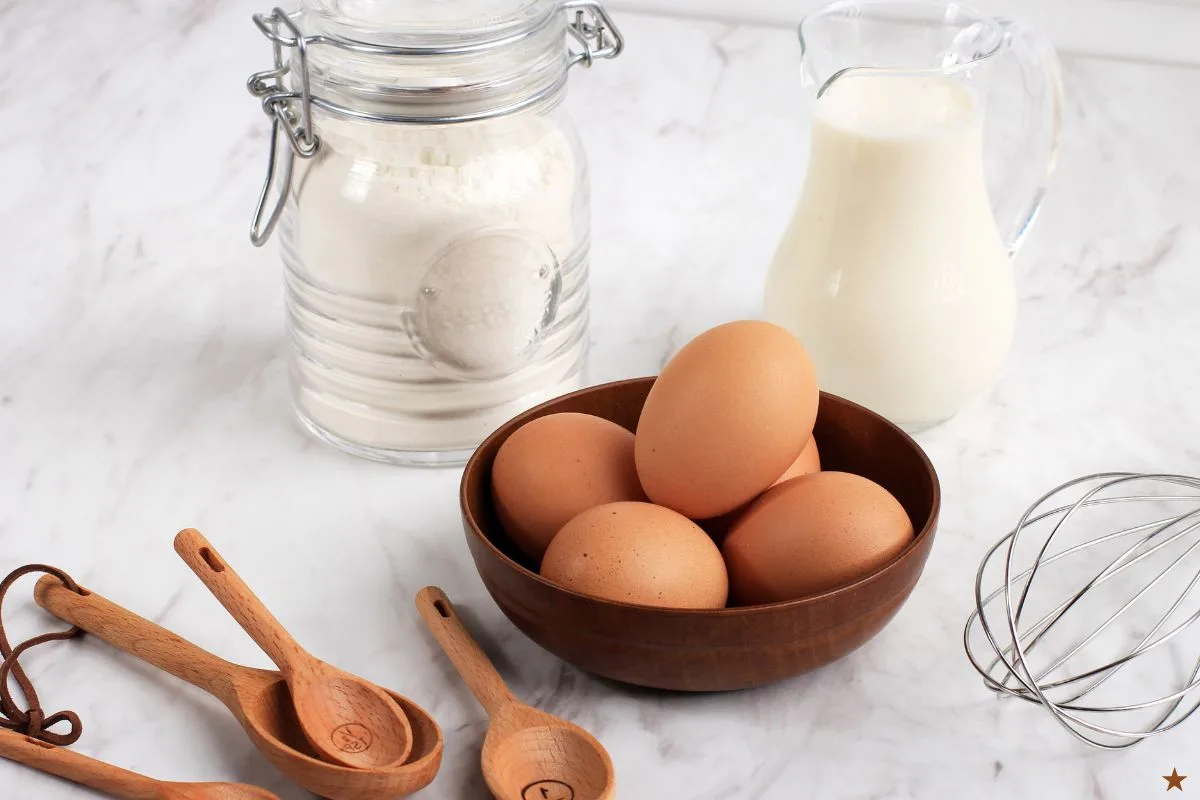 ingredientes para o bolinho de chuva assado: ovos, leite farinha em cima de uma superfície com fuê e colheres medidoras ao lado.