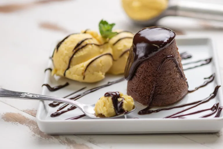 receita de petit gateau: bolinho de chocolate em um prato de sobremesa ao lado de duas bolas de sorvete e uma colher.
